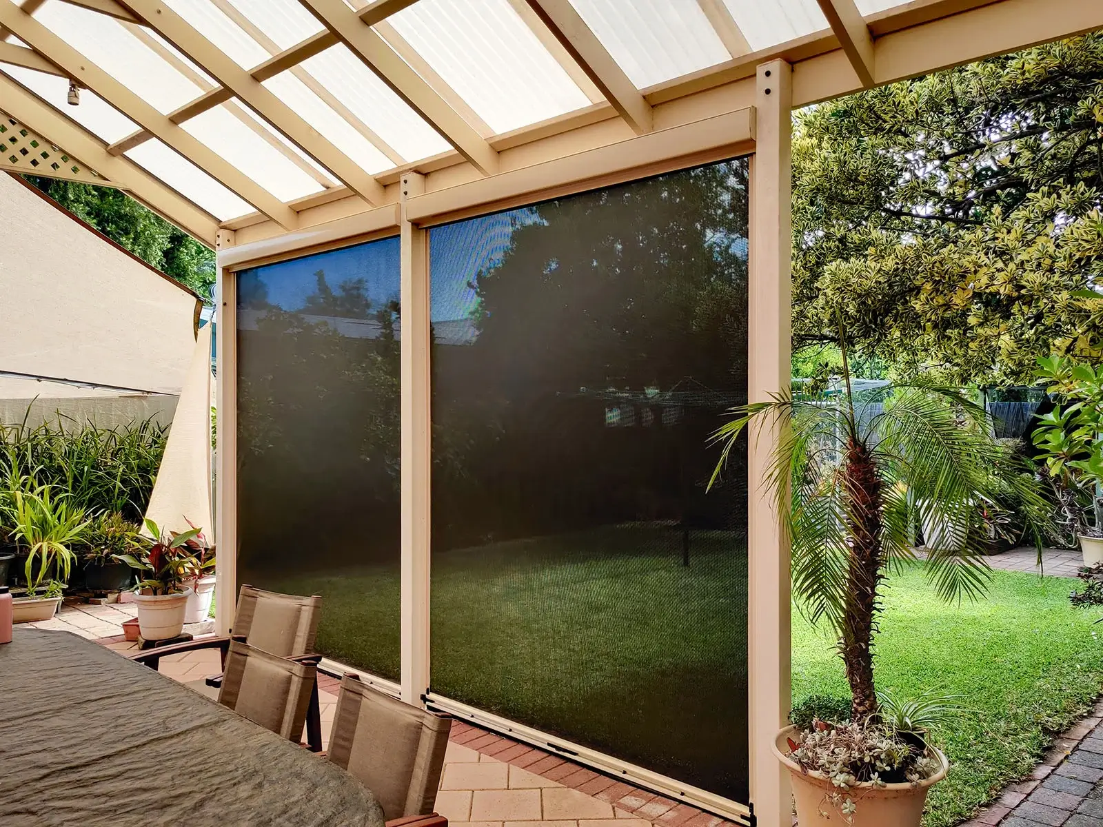 Outdoor dining area with Manual Ziptrak Blinds, providing shade and privacy, surrounded by potted plants and a lush green garden.